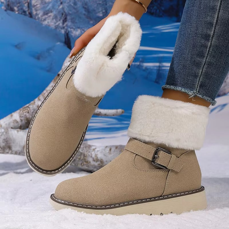 A person wearing beige ankle winter boots with thick white faux-fur trim and a buckle strap, standing in snow with a snowy mountain backdrop.