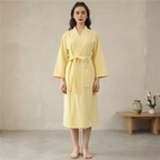 Woman wearing a yellow robe standing in a room with a wooden table and vase.