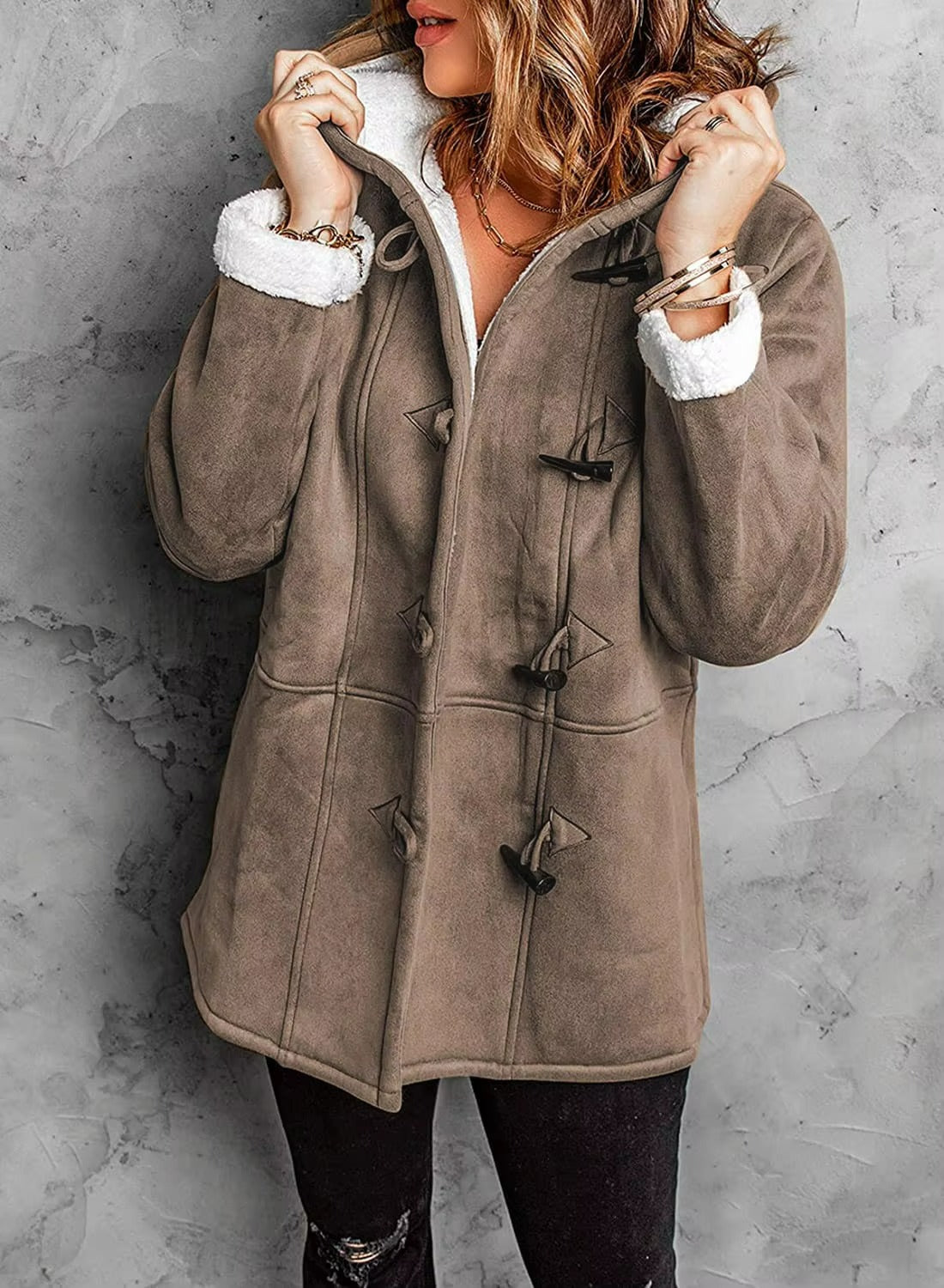 Person wearing a brown duffel coat with white lining against a gray concrete wall.