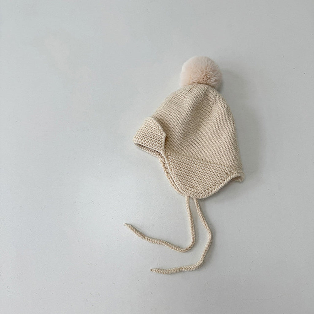 A cream-colored knit baby bonnet with a matching pom-pom and long tie strings, photographed on a neutral light gray background.