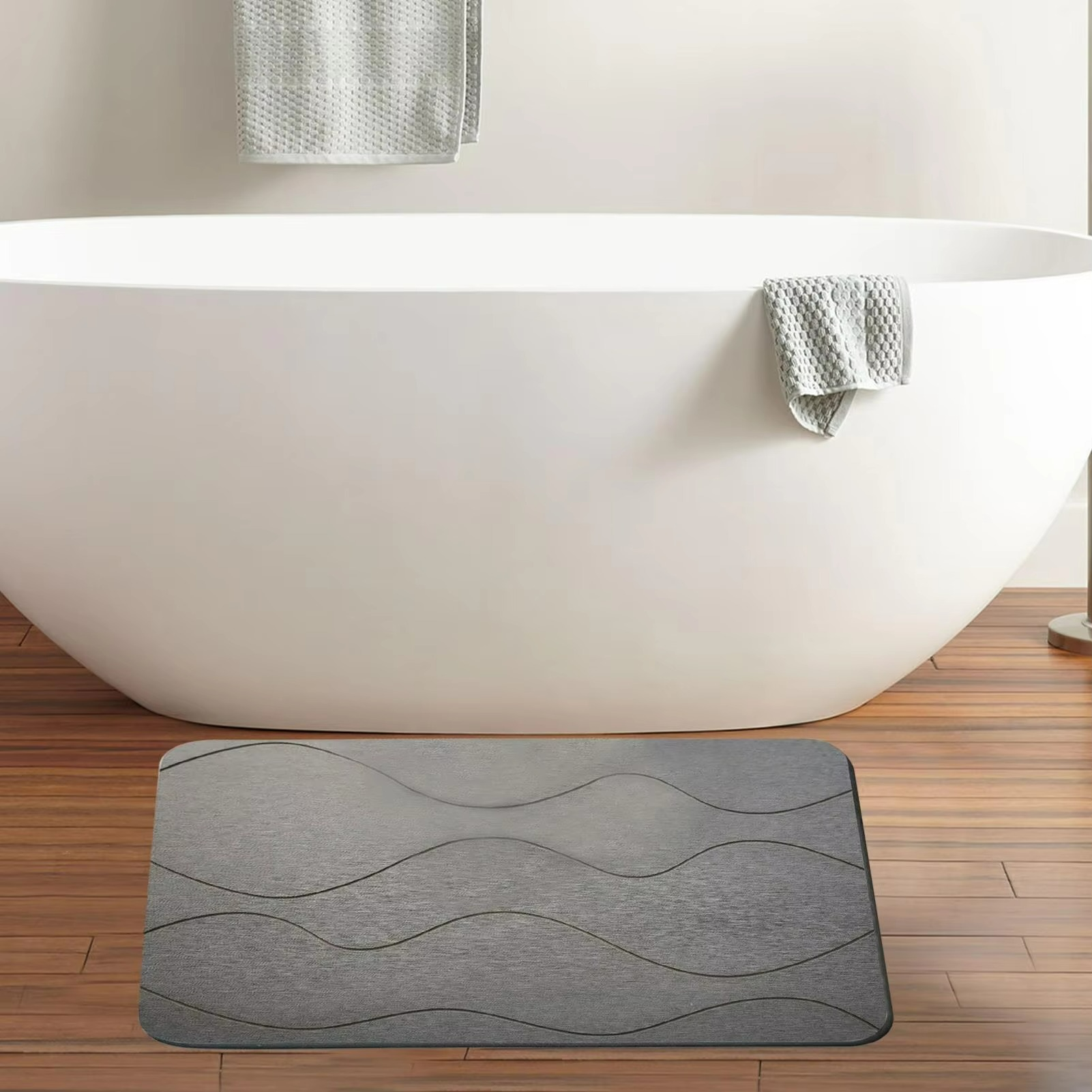 Gray textured bath mat in front of a white bathtub with towels on a wooden floor.