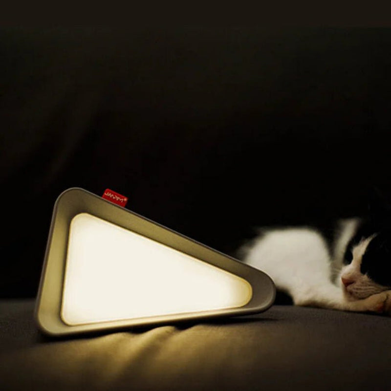 A triangular LED lamp with a glowing warm light and a small red brand tab, placed on a dark surface next to a sleeping black-and-white cat.