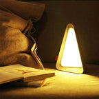 Triangle-shaped night light on a surface next to a book in a dimly lit room.