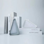 A minimalist gray triangular wall sconce with a frosted glass shade, styled on white display blocks next to a matching white triangular tray against a light gray background.