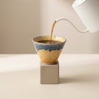 Ceramic coffee filter with blue rim being filled with coffee from a white kettle on a beige surface.