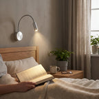 Person reading a book in bed with a night lamp on the wall.
