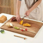 Person slicing oranges on a wooden cutting board with various fruits and vegetables.