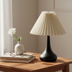 Table lamp with pleated shade on a wooden table next to books and a vase with a flower.