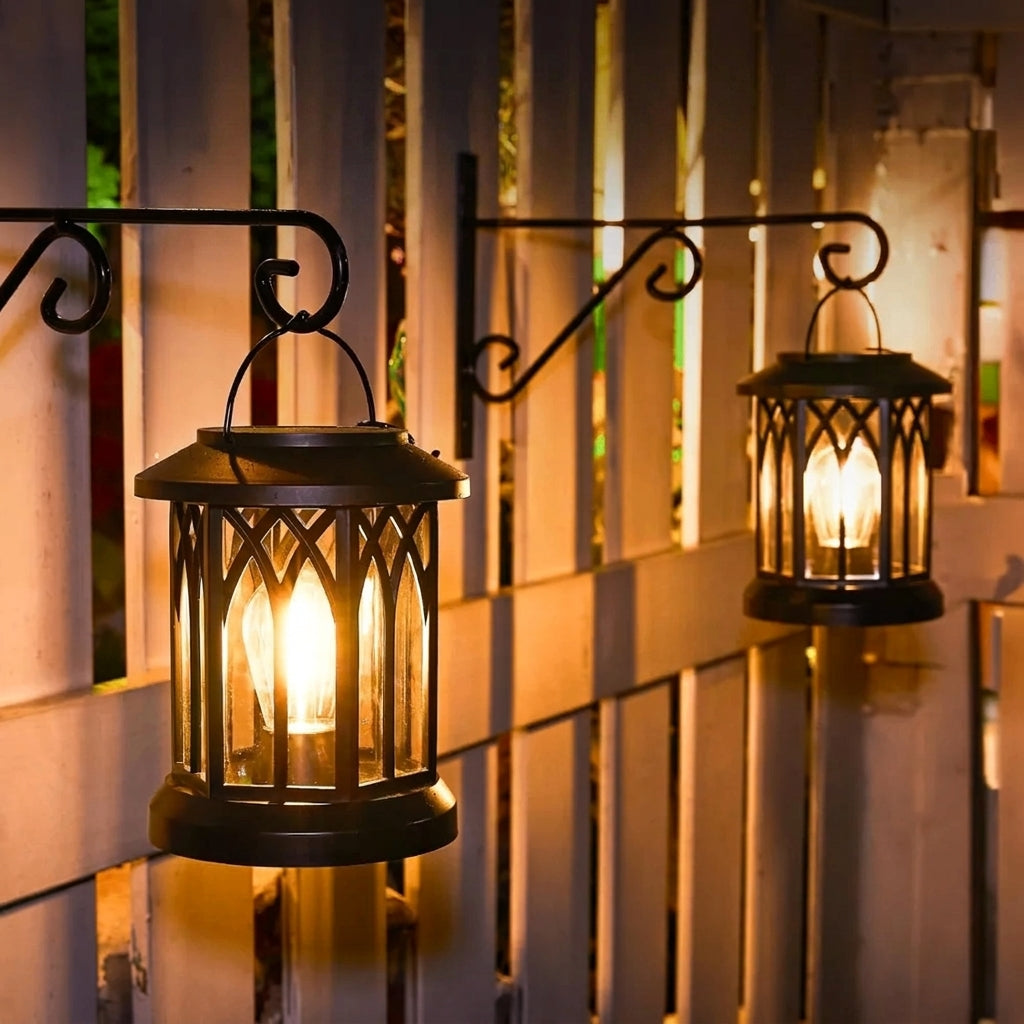 Two lit outdoor lanterns hanging against a wooden fence during sunset.