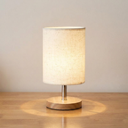 A table lamp with a cylindrical beige linen shade, a slim metallic stem, and a round light wood base, shown lit on a wooden tabletop against a plain light wall.