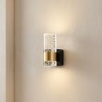 A modern wall-mounted light fixture with a clear cylindrical glass shade, a brushed gold middle band, and a black backplate, shown lit on a beige wall.