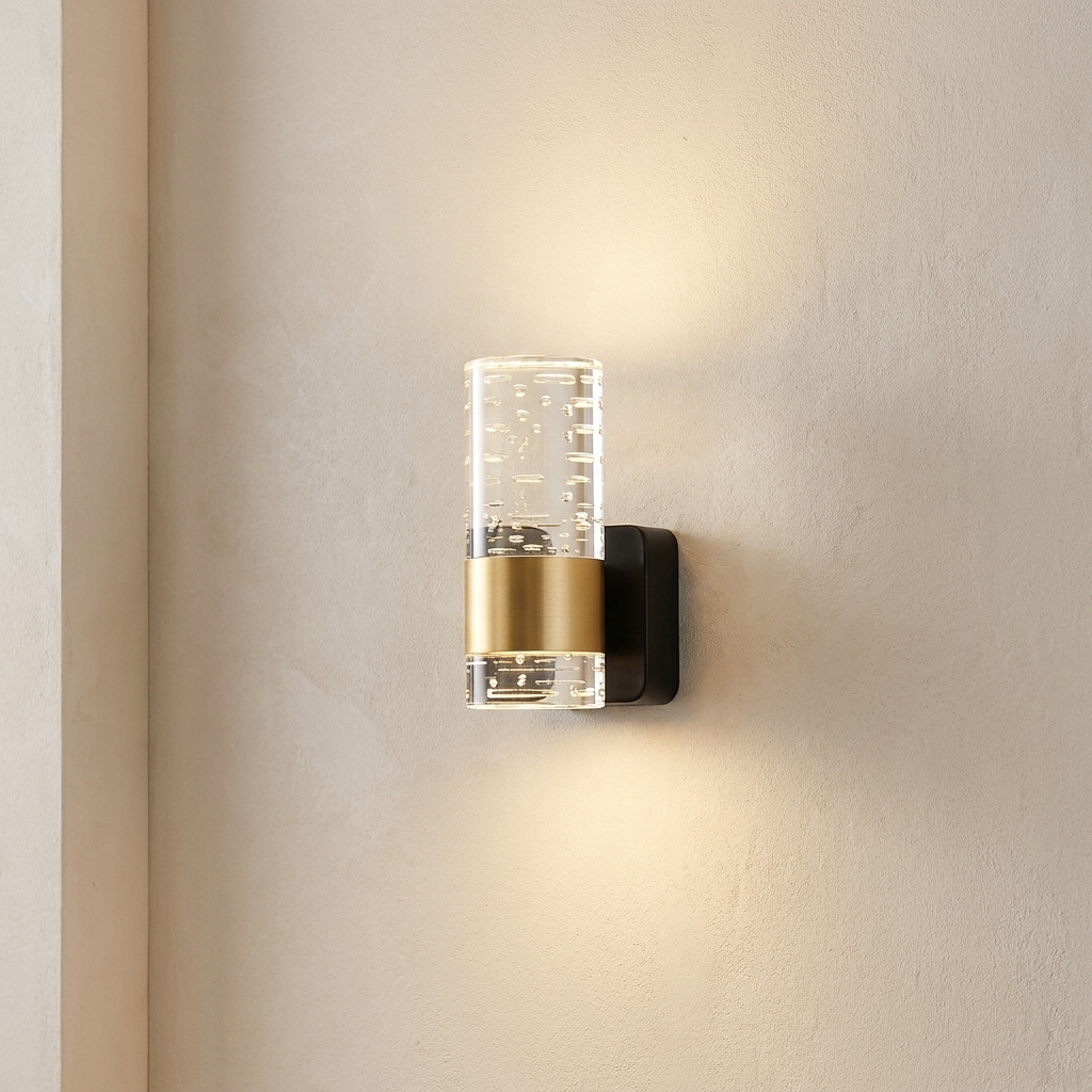 A modern wall-mounted light fixture with a clear cylindrical glass shade, a brushed gold middle band, and a black backplate, shown lit on a beige wall.