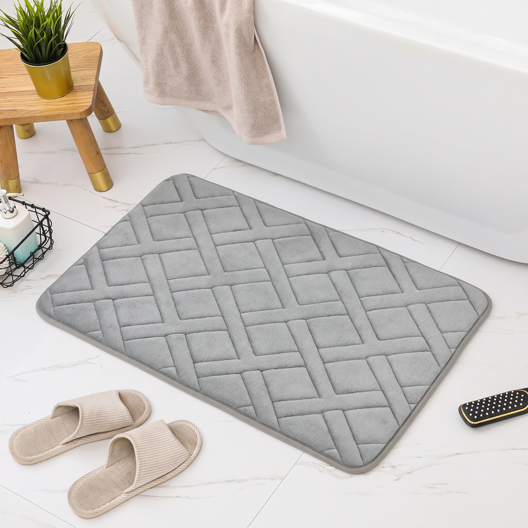 A gray rectangular bath mat with a raised basketweave pattern, styled in a bathroom setting with a white bathtub, beige slippers, a towel, and a small wooden stool.