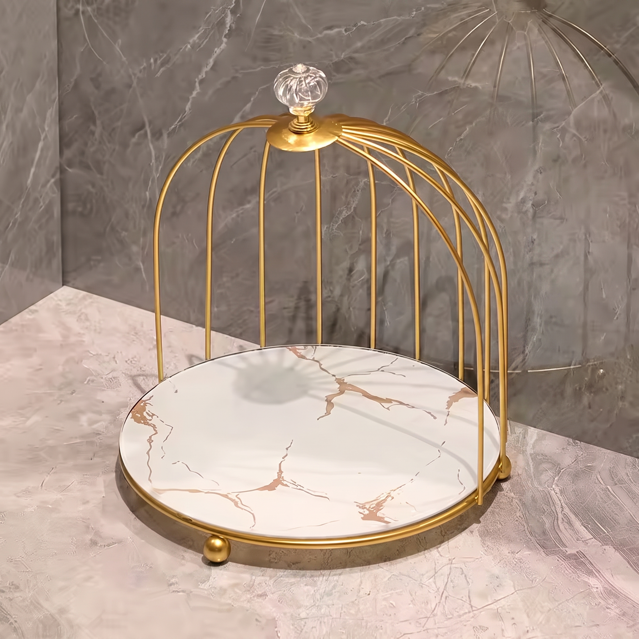 A decorative gold metal birdcage-style stand with a round white marble-patterned tray base, shown on a marble surface against a gray marble wall.