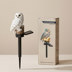A decorative owl garden stake with a white owl figurine perched on a small wooden perch and black stake, shown beside its illustrated product box on a clean beige studio background.