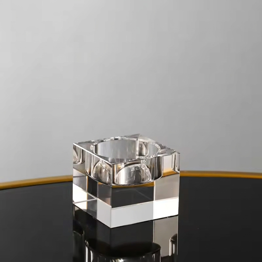 A clear glass or acrylic square ashtray with a reflective, polished finish, standing on a black reflective surface against a light gray background.