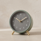 A minimalist round alarm clock with a sage green metal body, a dark gray face, and gold-toned hands and feet, set against a neutral beige background.