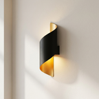 A modern wall-mounted light fixture with a black outer shell and a gold inner panel, shown lit against a warm off-white wall.
