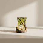 A small decorative glass vessel with a sculptural, wavy form, featuring an embedded tree design in yellow-green tones and a natural wood base, set against a minimalist beige background with soft diagonal shadows.