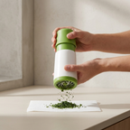 A person's hands holding a green and white manual herb grinder over a white paper towel on a beige countertop, with chopped green herbs falling out.