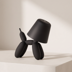 A matte black balloon dog table lamp shaped like a dog with a conical lampshade head, standing on a white pedestal against a warm beige background.
