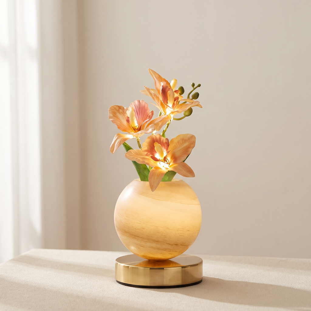 A decorative lamp-shaped vase with a glowing warm light, featuring peach-orange artificial flowers and green stems, set on a round metallic base against a soft beige background.