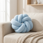 A light blue knotted round throw pillow resting on a beige fabric sofa with a textured cream throw blanket in a softly lit room.
