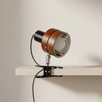 A modern table lamp with a round wooden shade, black metal arm, and clear glass lens, mounted on a white shelf against a beige wall.
