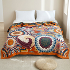 A colorful patterned comforter or duvet cover spread across a bed, featuring large circular and spiral motifs in orange, blue, cream, burgundy, and yellow. The bed is styled with two white pillows and white bedding, set in a bright, neutral bedroom with a woven floor cushion in front.
