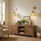 Two matching wall-mounted sconces with green ribbed glass shades and brass fixtures are shown lit in a styled room above a wooden console cabinet, with a chair, rug, books, and framed art visible.