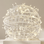 A spherical decorative sculpture made of intertwined white branches or wire, illuminated with many small warm white string lights, displayed on a white pedestal against a soft beige background.