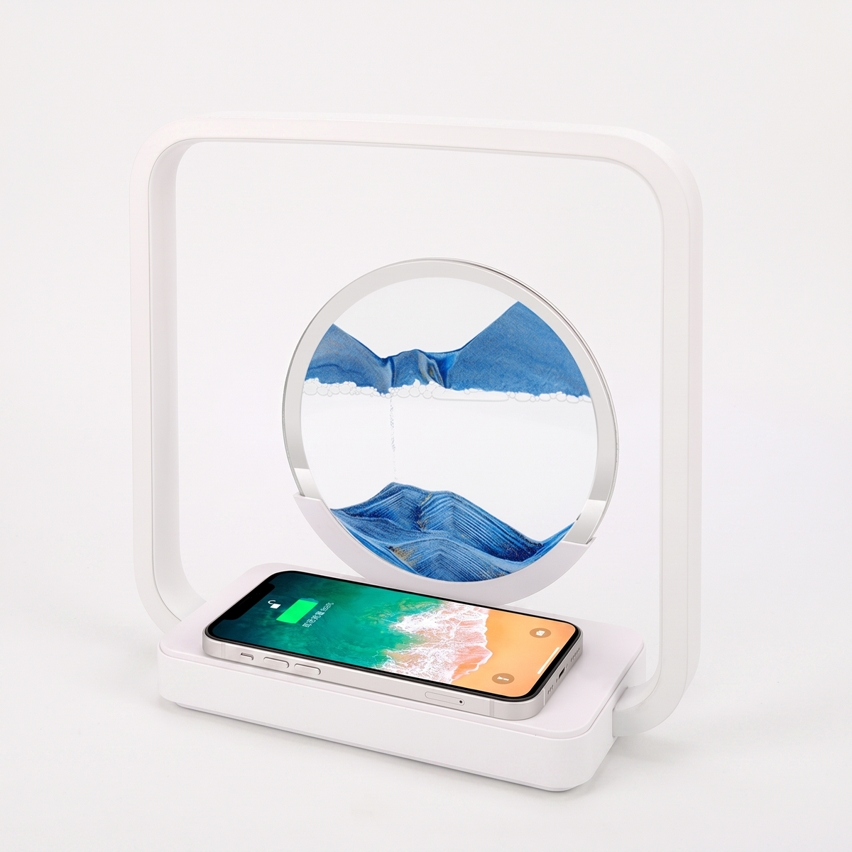 A white rectangular wireless charging stand with a circular mirror on top, shown on a clean white background. A smartphone lies on the charging base, and the mirror reflects a blue mountain-and-sea scene.