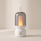 A modern white tabletop lamp with a translucent smoky glass dome and a glowing warm yellow candle-style bulb inside, shown against a soft beige background.