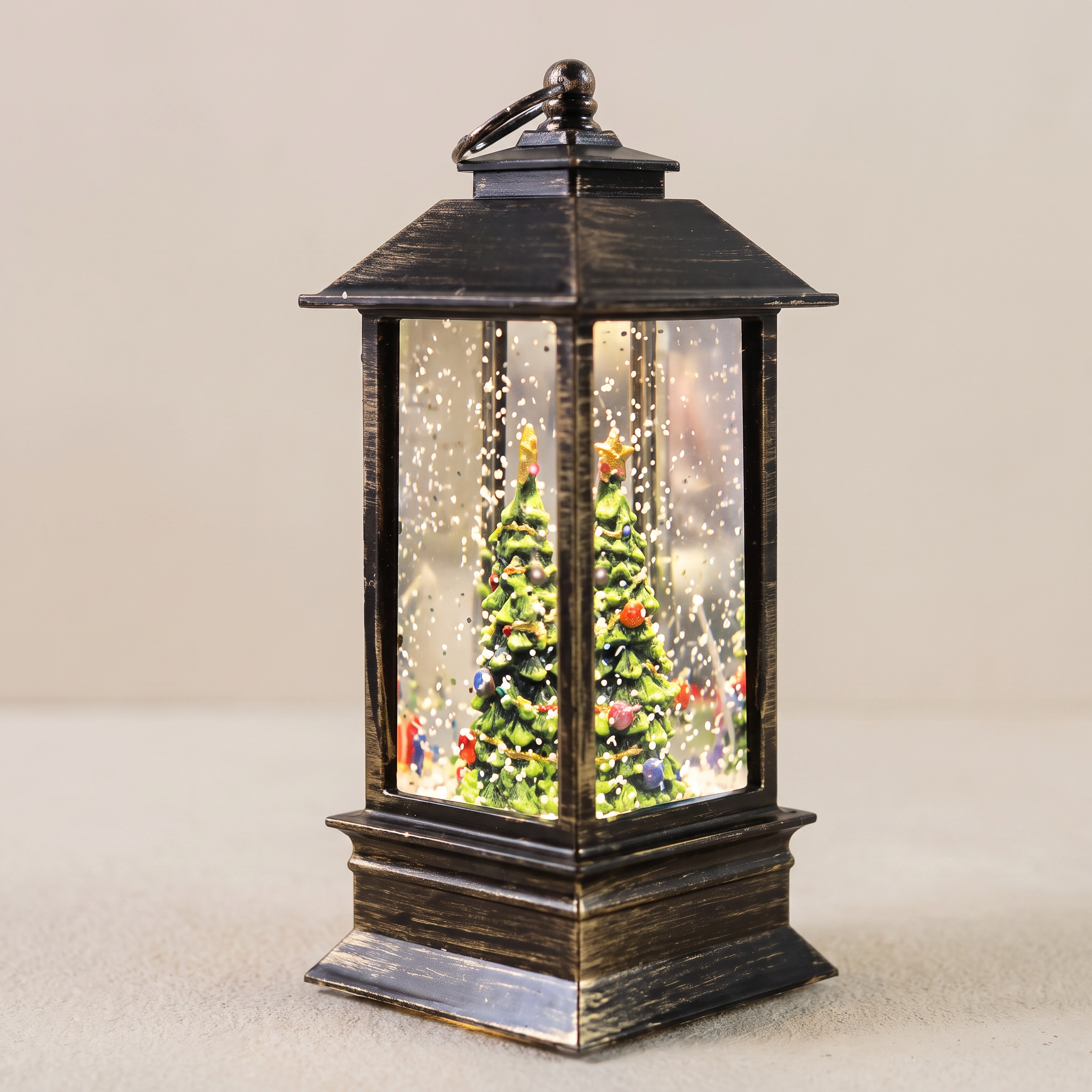 A decorative black lantern with a miniature Christmas tree inside, featuring falling snowflake details and colorful ornaments, set against a plain beige background.