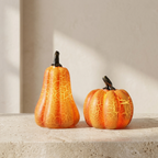 Two decorative faux pumpkins with a crackled orange and yellow finish and dark stems, displayed on a light stone surface against a warm beige background.