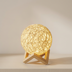 A spherical woven lamp with a warm glowing light, resting on a light wood geometric stand against a soft beige background.