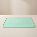 A mint green silicone baking mat with concentric circle guides and measurement markings, resting on a light beige surface against a matching background.