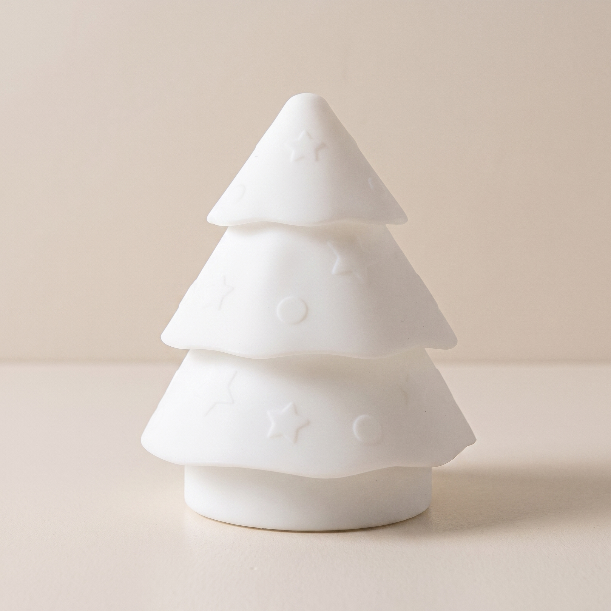 A small white Christmas tree-shaped decorative object with embossed star and circle patterns, standing on a matching round base against a warm beige background.