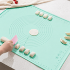 A mint green silicone dough rolling mat with measurement markings and concentric circles, shown on a light countertop. A hand is using a pink scraper to handle dough pieces, with several small dough balls and dumplings arranged on the mat.