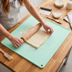 A mint green silicone baking mat with measurement markings is shown on a wooden countertop, with a person rolling out dough using a wooden rolling pin. The scene includes baking tools like a brush, scraper, and flour containers.