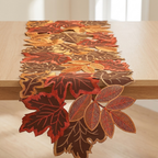 An autumn-themed table runner made of layered embroidered leaf cutouts in shades of red, orange, brown, and gold, draped over a light wood table.