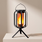 A black metal lantern table lamp with a glowing orange and yellow faux flame inside a clear glass cylinder, standing on a tripod base against a warm beige background.