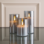 Three metallic silver cylindrical candle holders with lit flameless candles, arranged on a reflective dark surface against a warm beige paneled background.