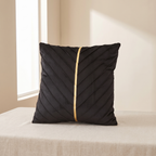A square black throw pillow with diagonal pleated stitching and a central gold stripe, resting on a beige fabric-covered surface in a softly lit room.