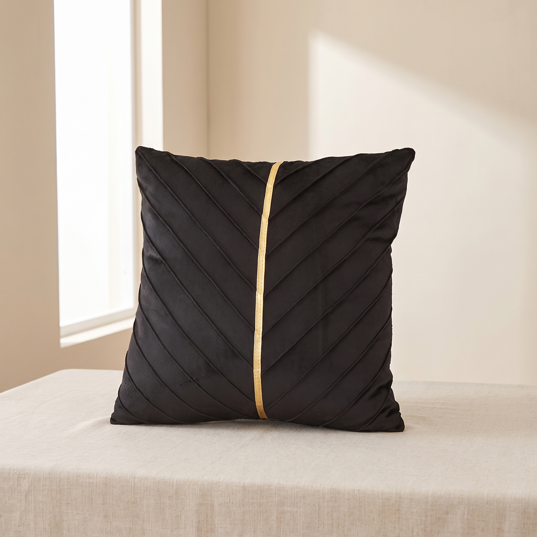 A square black throw pillow with diagonal pleated stitching and a central gold stripe, resting on a beige fabric-covered surface in a softly lit room.