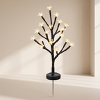 A black solar-powered decorative tree light with small glowing white flower bulbs on branching limbs, mounted on a stake with a visible circular solar panel base.