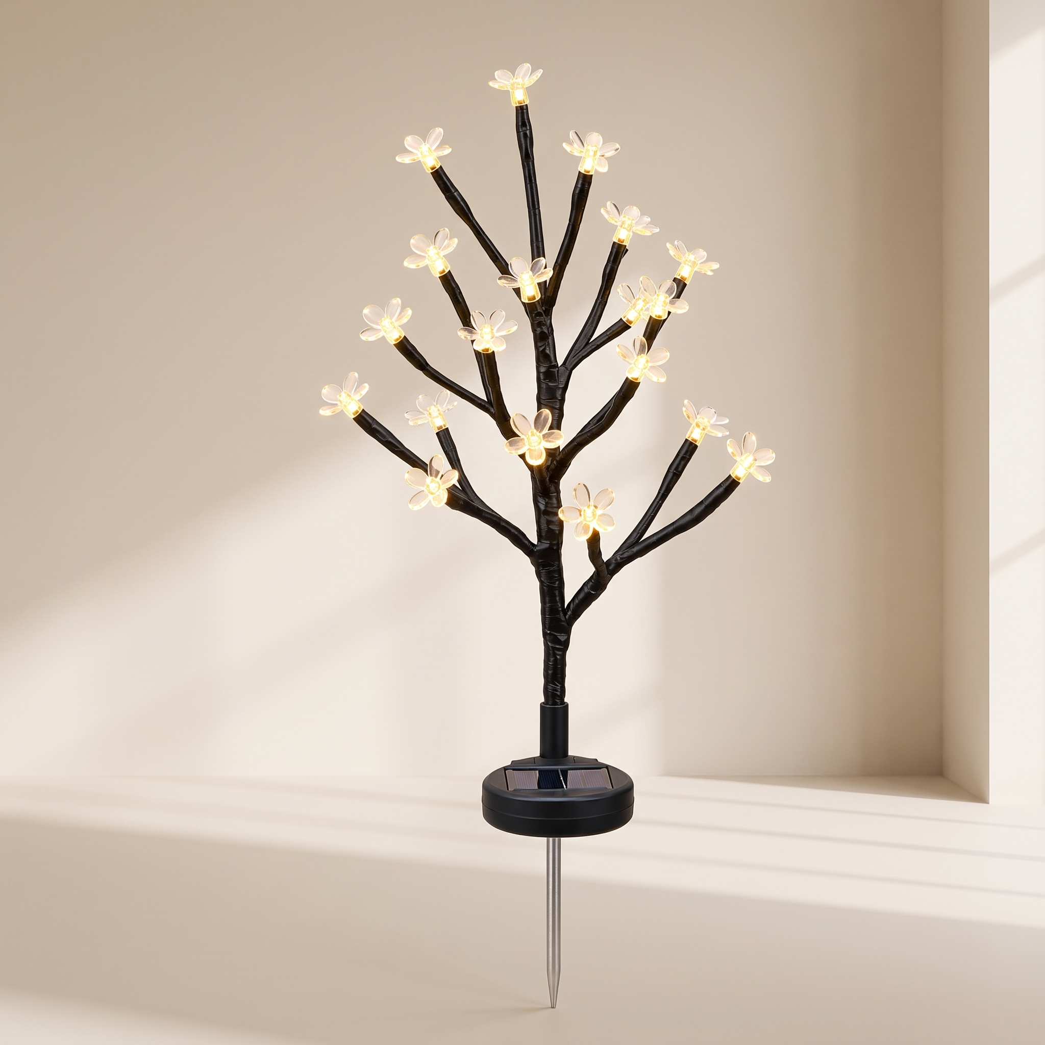 A black solar-powered decorative tree light with small glowing white flower bulbs on branching limbs, mounted on a stake with a visible circular solar panel base.
