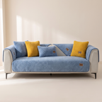 A blue sofa slipcover fitted over a light gray sofa, styled with four throw pillows in blue and mustard yellow, set against a warm beige wall with soft window shadows.