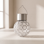 A clear glass solar lantern with a textured bubble pattern, a brushed silver metal cap, and a wire handle, set on a light beige surface near a window.