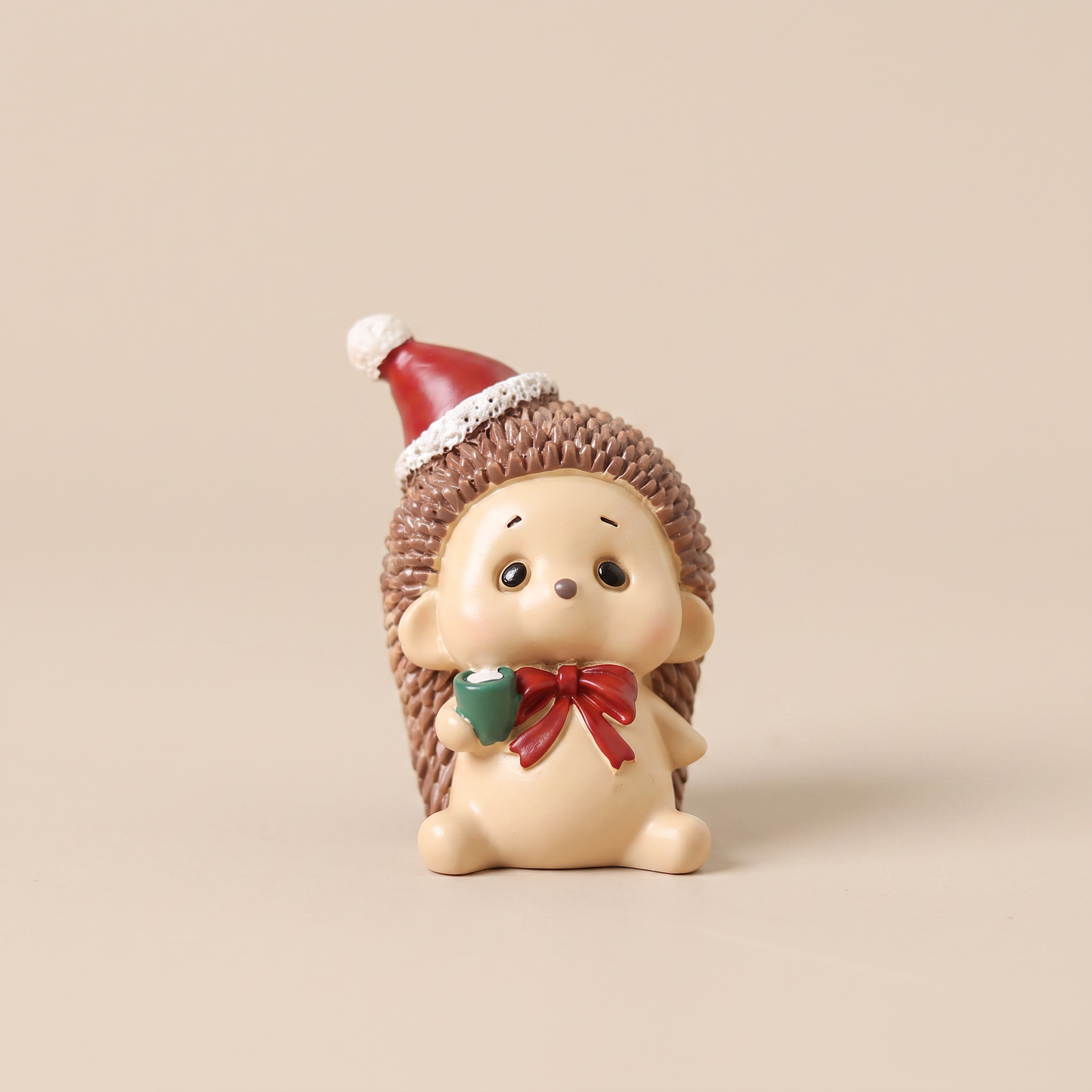 A small decorative hedgehog figurine with a cream-colored face and body, brown spiky texture, a red Santa hat, and a red bow with a green accent, standing against a plain beige background.